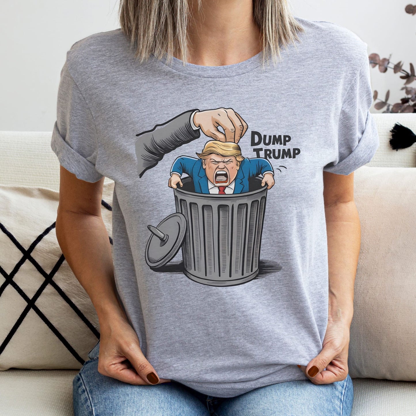 Person wearing a gray t-shirt with a graphic of a hand dumping a figure labeled 'Dump Trump' into a trash bin: Anti Trump shirt, Anti Trumpism shirt, Dump Donald Trump US election 2024 tshirt, Pro Kamala Harris Walz tee, Funny political apparel