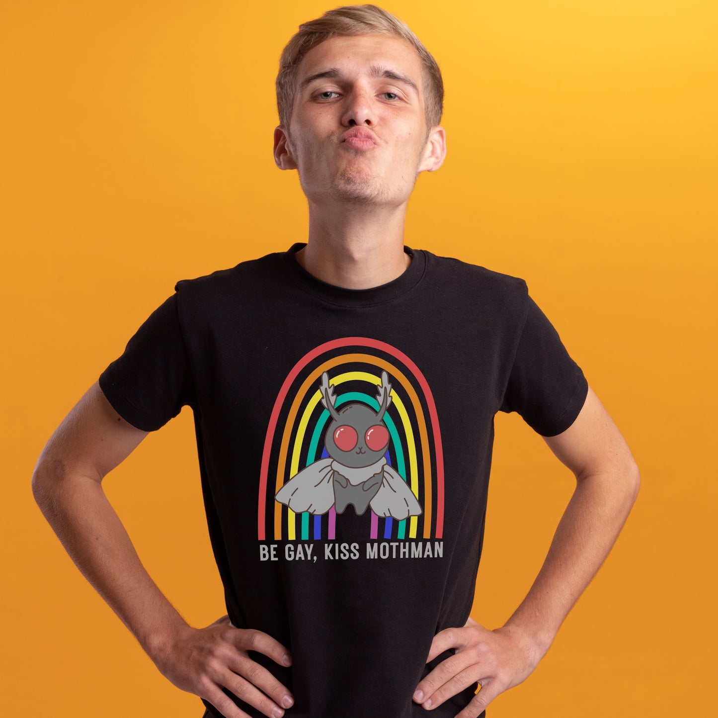 Person wearing a black t-shirt with a rainbow graphic and text on an orange background: LGBTQ Mothman Comfort Colors Shirt, Be Gay Kiss Mothman Tee, Queer Cryptid T-Shirt, LGBT Cryptidcore Gift, Rainbow Cryptozoology Merch