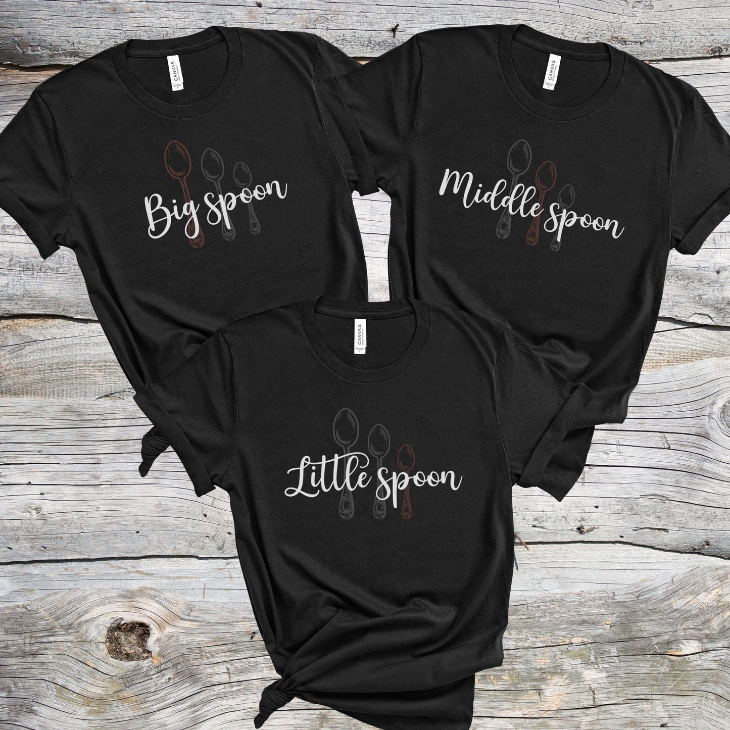 Three black matching ENM polyamory t-shirts with 'Big spoon', 'Middle spoon', and 'Little spoon' text on a wooden surface.