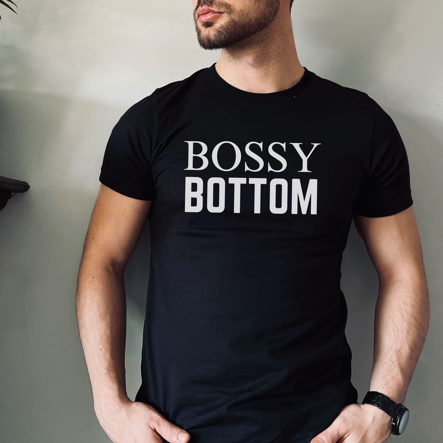 Queer person wearing a black t-shirt with 'BOSSY BOTTOM' text against a neutral background