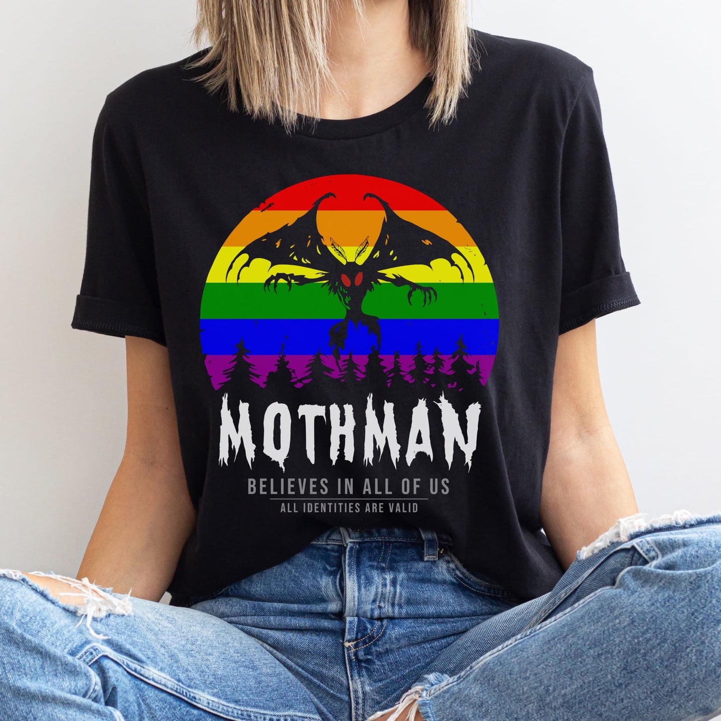 Person wearing a black t-shirt with a rainbow Mothman graphic and text: LGBTQ Mothman Shirt, Rainbow Pride Cryptid Folklore T-Shirt, Queer Mythology Legend Cryptozoology Tee, Cryptidcore Weirdcore Gift For LGBT