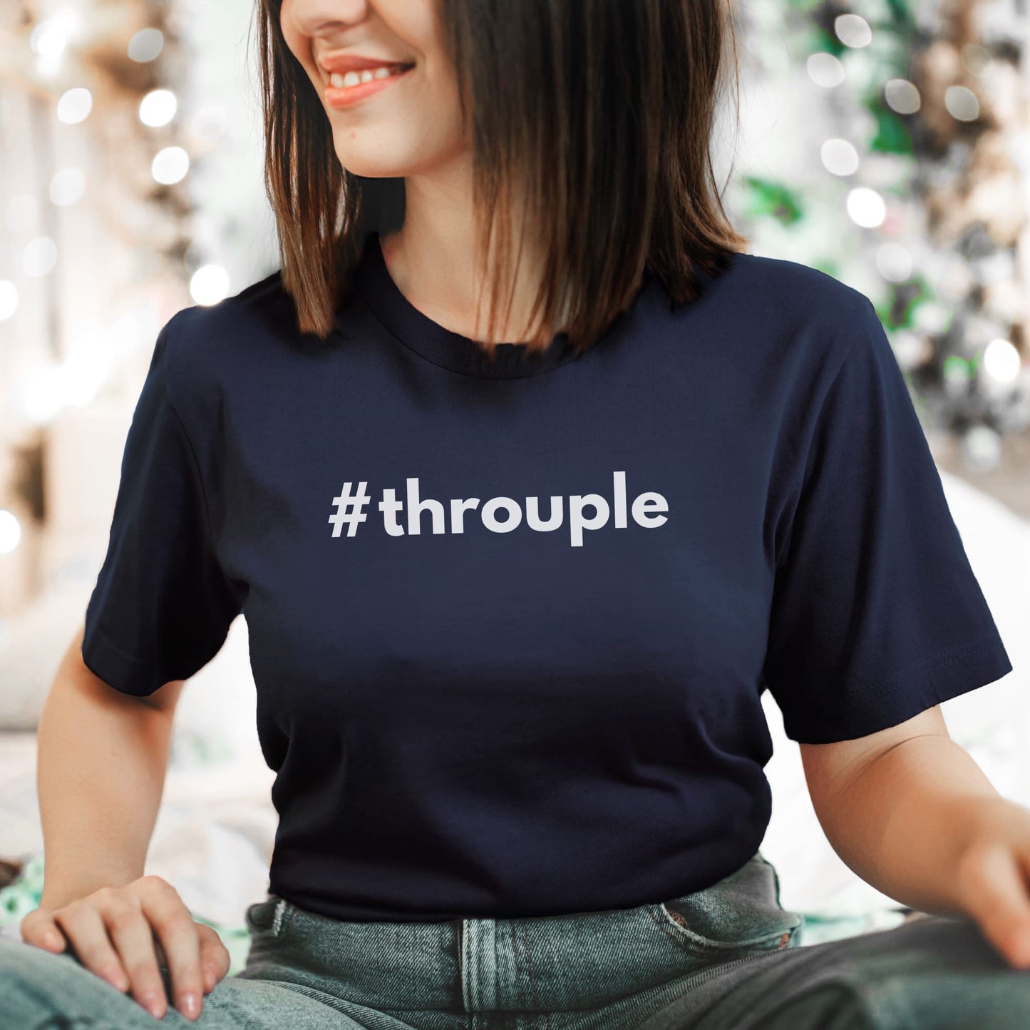Person wearing a navy blue t-shirt with '#throuple' ENM polyamory text, sitting against a blurred indoor background.