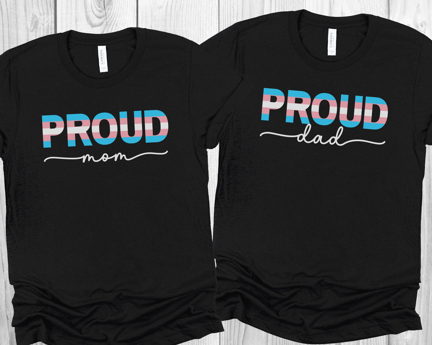 Two black transgender supporting t-shirts with 'PROUD mom' and 'PROUD dad' text on a wooden surface.
