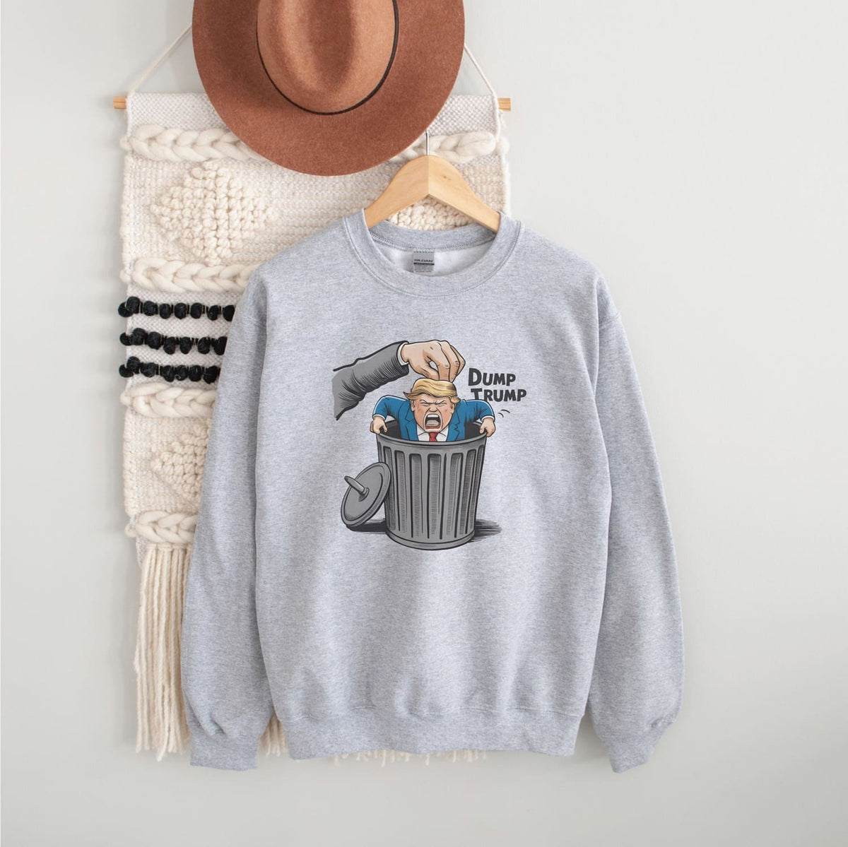 Gray sweatshirt with 'Dump Trump' graphic on a hanger against a white wall.