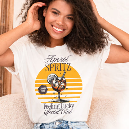 Woman wearing a white t-shirt with a drink graphic and text, sitting on a couch: Aperol Spritz Social Club Shirt, Summer Day Drinking Tee, Cocktail Party Girls Night Out T-Shirt, Aperol Lover Gift, Bartender Outfit
