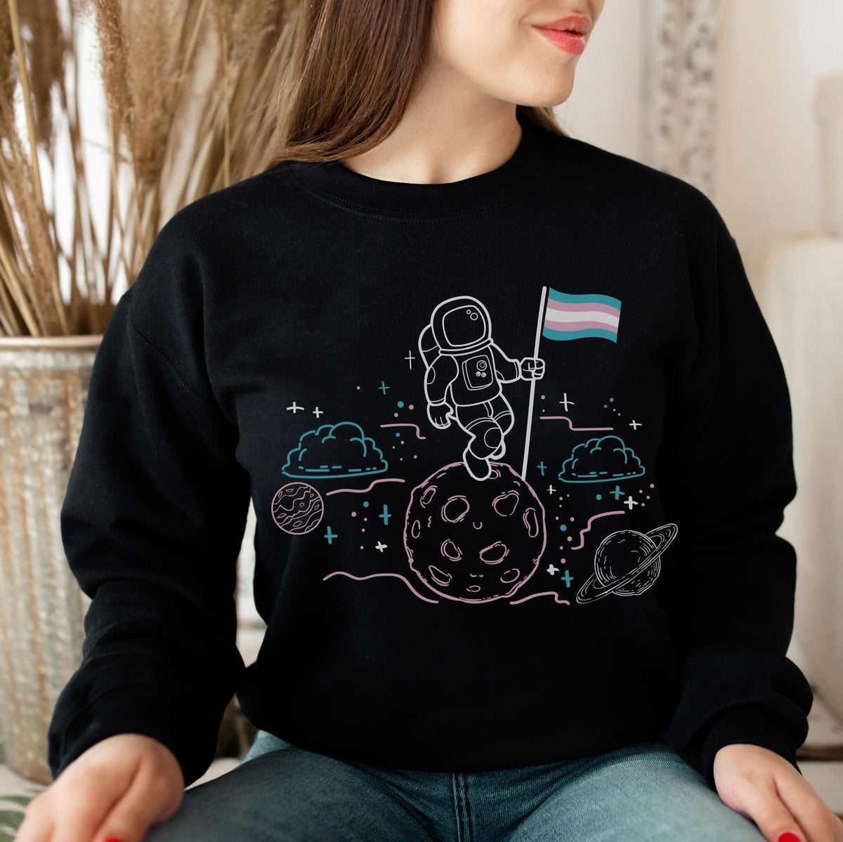 Person wearing a black sweatshirt with an astronaut transgender design on a moon surface, holding a trans flag.