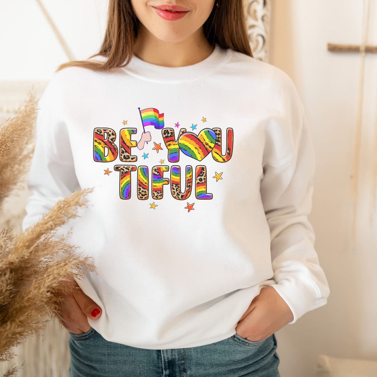 Person wearing a white pride sweatshirt with 'BE YOU TIFUL' text in rainbow colors.