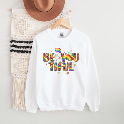 White pride sweatshirt with rainbow lgbtq 'BE YOU TIFUL' text on a hanger with a hat and scarf in the background.