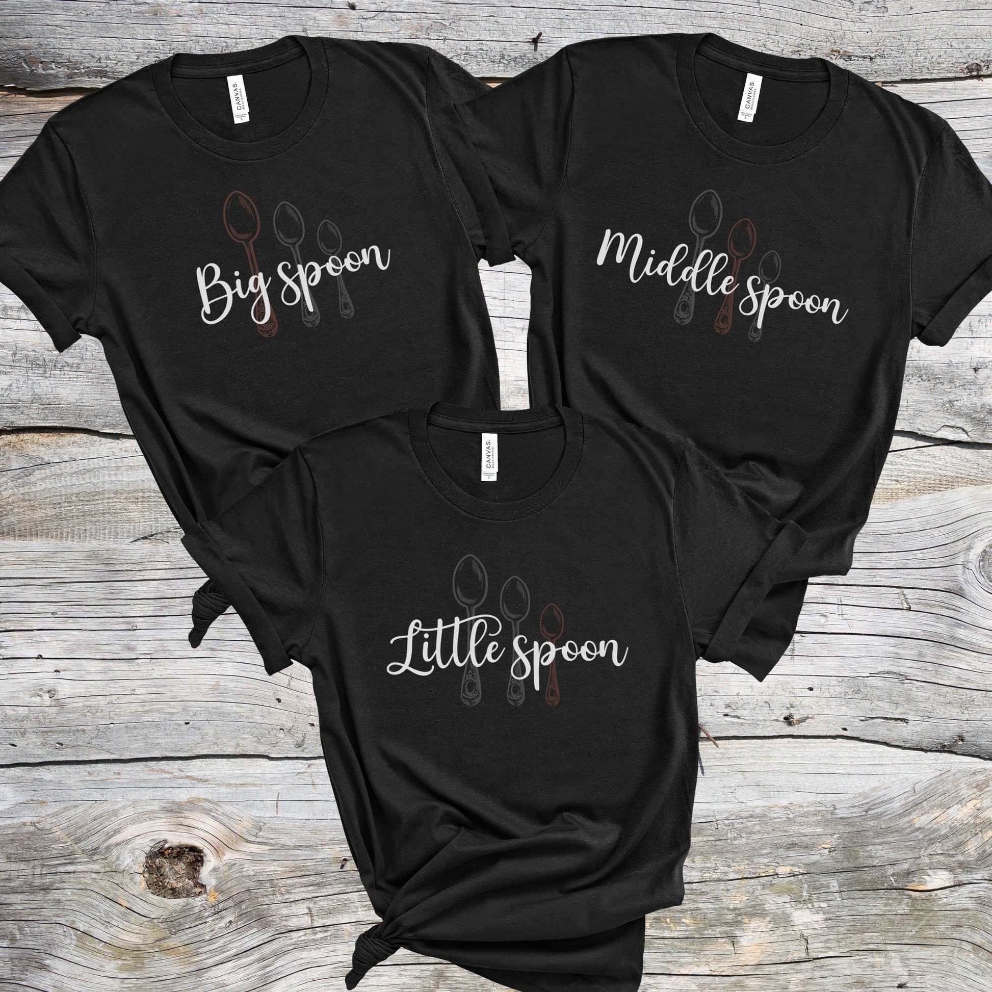Three black matching ENM polyamory t-shirts with 'Big spoon', 'Middle spoon', and 'Little spoon' text on a wooden surface.