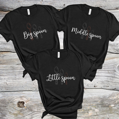 Three black matching ENM polyamory t-shirts with 'Big spoon', 'Middle spoon', and 'Little spoon' text on a wooden surface.