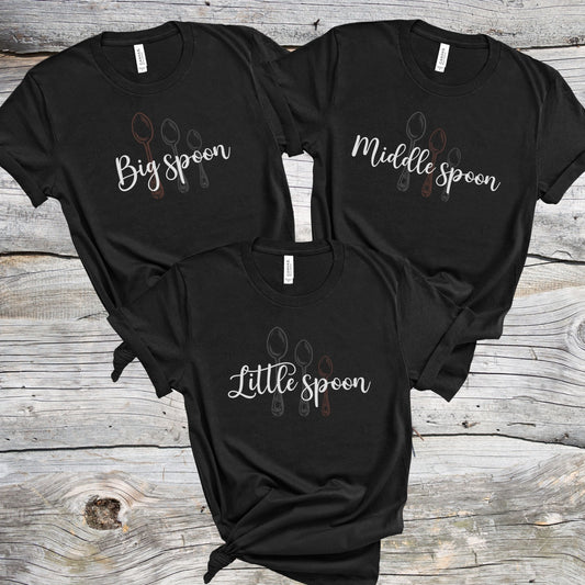 Three black matching ENM polyamory t-shirts with 'Big spoon', 'Middle spoon', and 'Little spoon' text on a wooden surface.