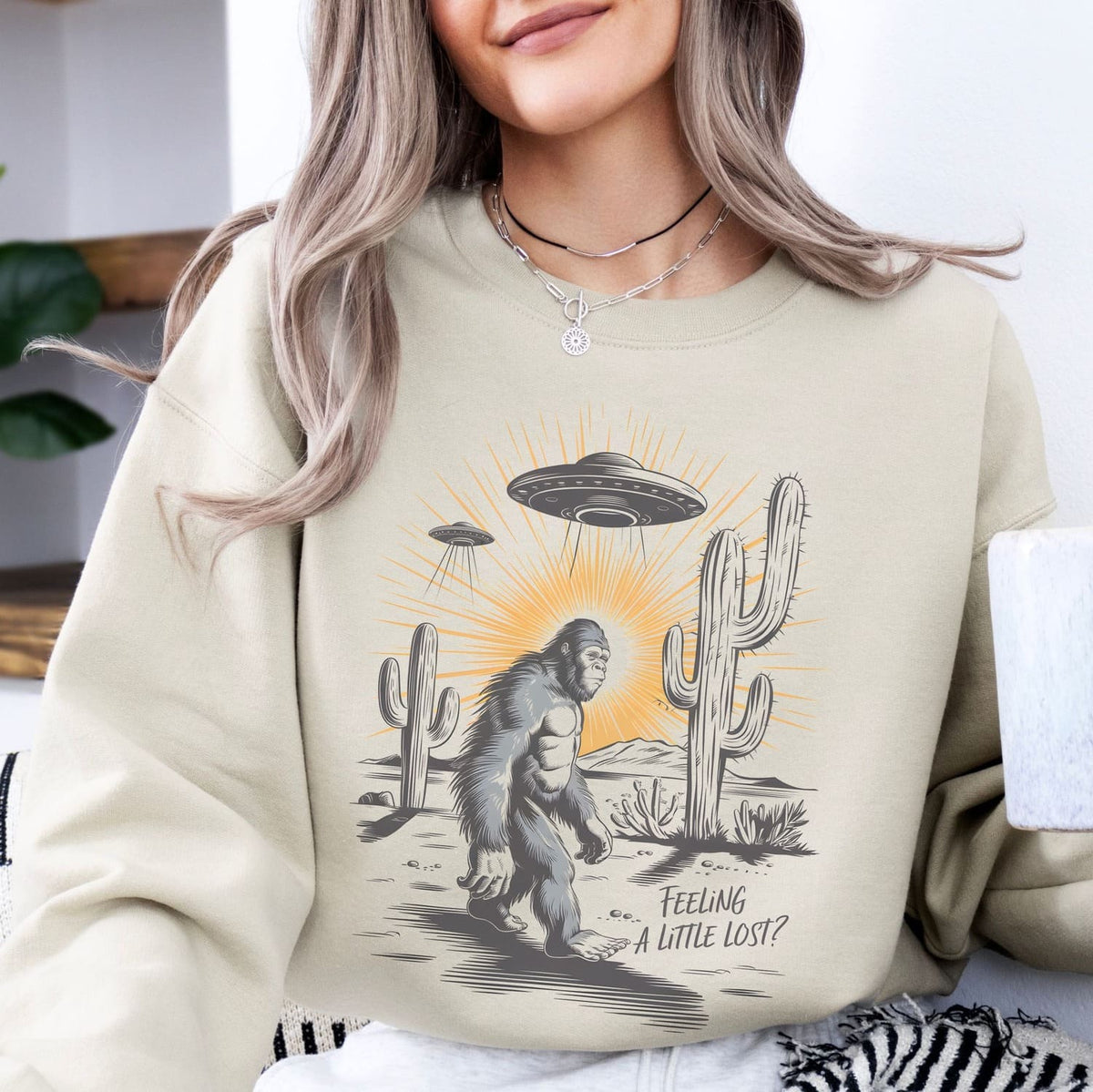 Person wearing a beige sweatshirt with a graphic of bigfoot, UFO, and cactus with text 'Feeling a little lost?'.