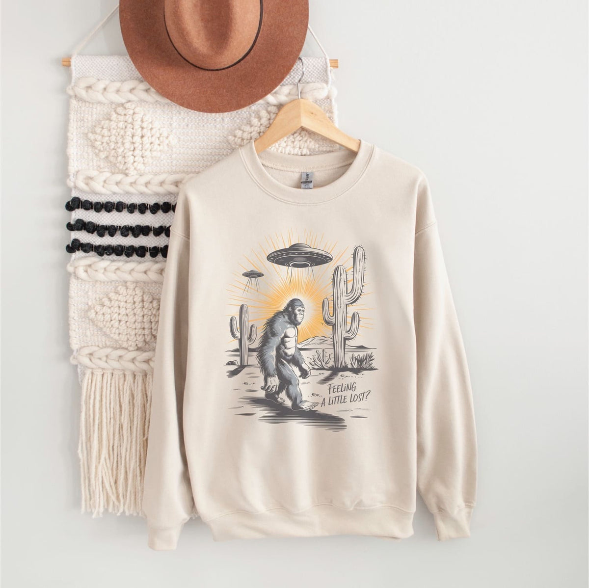Beige sweatshirt with graphic design of bigfoot, UFO, and cactus on a hanger against a white wall.
