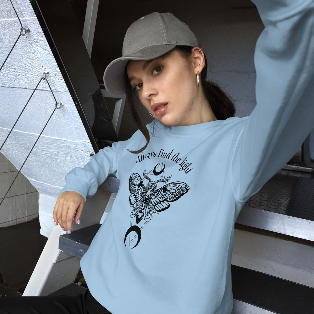 Person wearing a light blue inspirational sweatshirt with a black moth design and text, sitting on stairs.