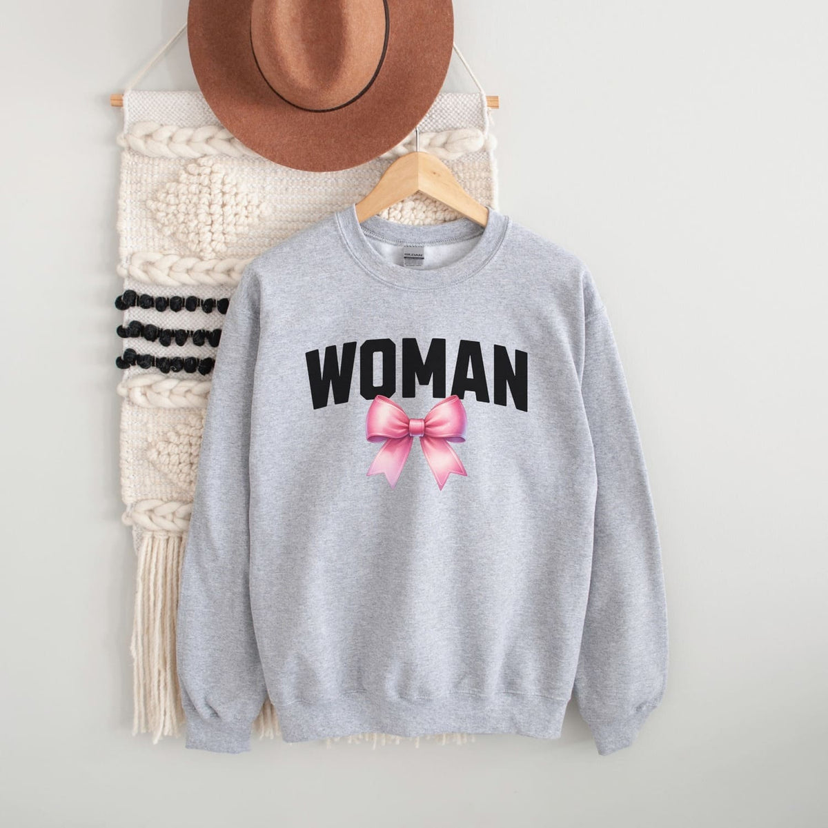 Gray mtf transgender sweatshirt with 'WOMAN' text and pink bow, hanging on a white wall with a hat and macramé wall art.