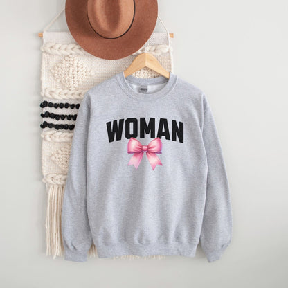 Gray mtf transgender sweatshirt with 'WOMAN' text and pink bow, hanging on a white wall with a hat and macramé wall art.