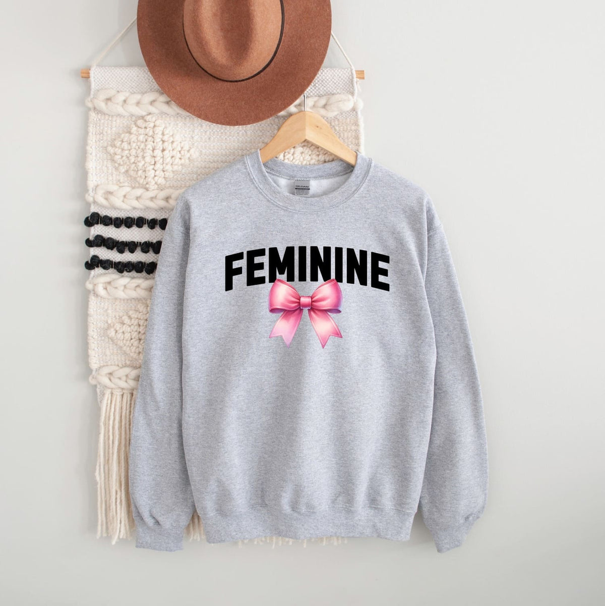 Gray sweatshirt with 'FEMININE' text and pink bow graphic, hanging on a white wall with a hat and macramé wall art.