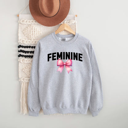 Gray sweatshirt with 'FEMININE' text and pink bow graphic, hanging on a white wall with a hat and macramé wall art.
