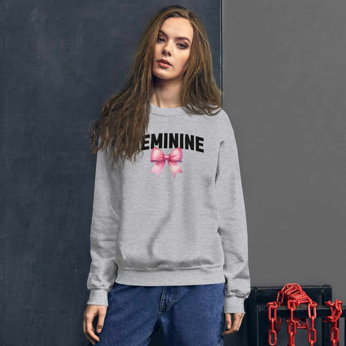 Woman wearing a gray sweatshirt with 'FEMININE' and a pink bow graphic, standing against a dark background.