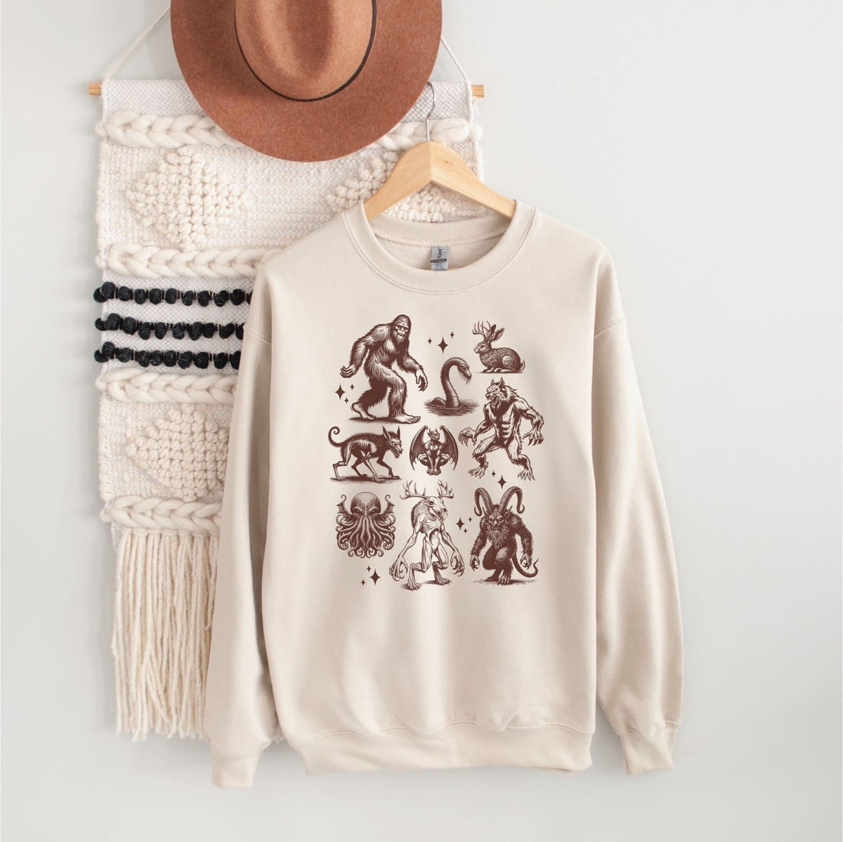 Beige sweatshirt with cryptids on a hanger against a white wall.