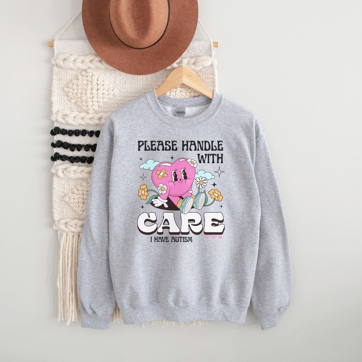 Gray autism sweatshirt with a heart design and text, placed on a white surface with a hat and bag in the background.