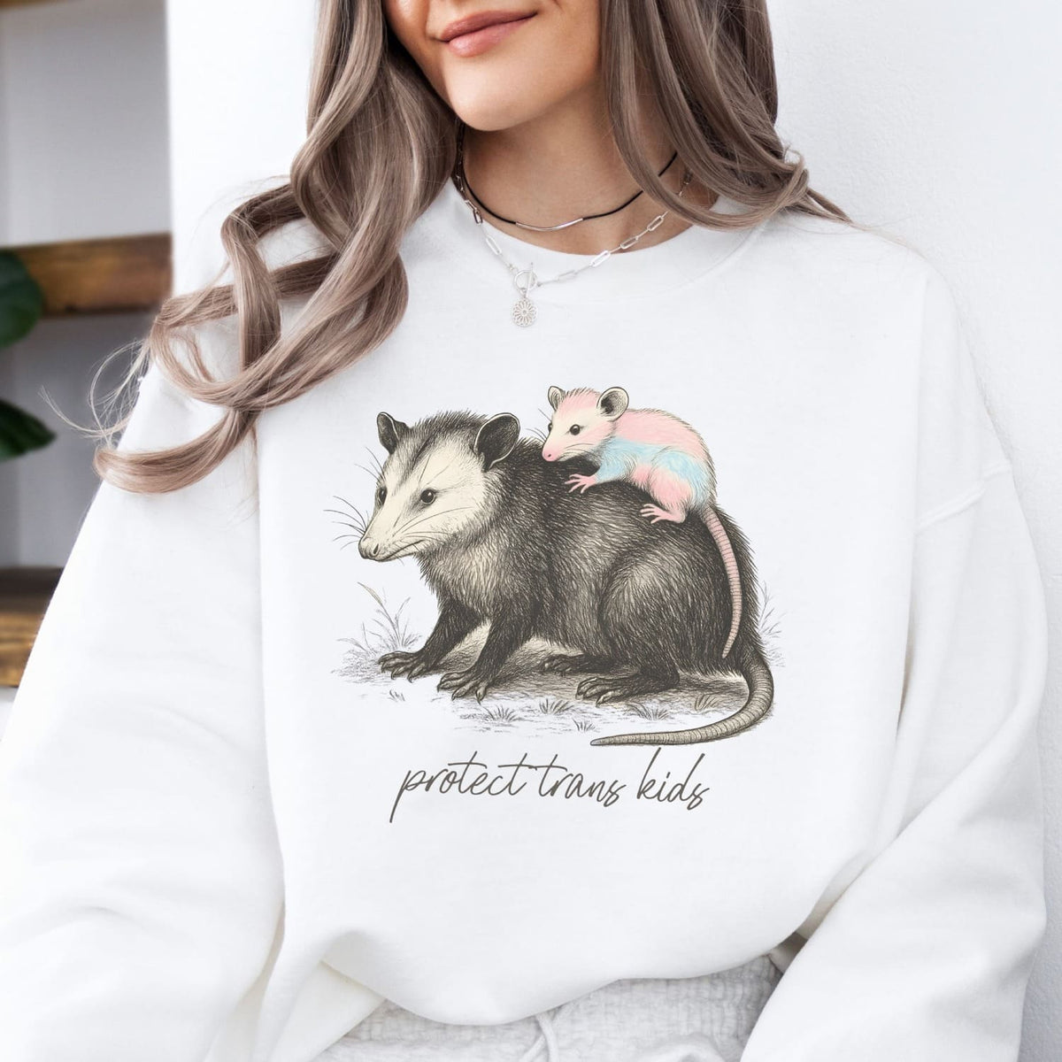 Person wearing a white sweatshirt with an illustration of an opossum and baby opossum, with text 'protect trans kids'.