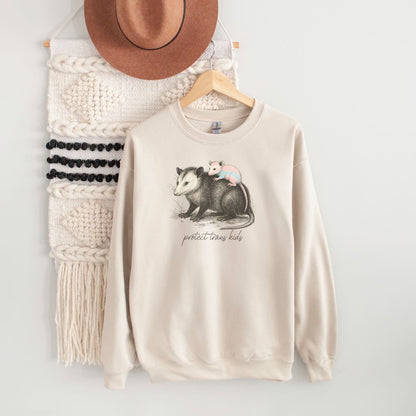 Beige 'protect trans kids' sweatshirt with opossum graphic and text on a hanger against a white wall.
