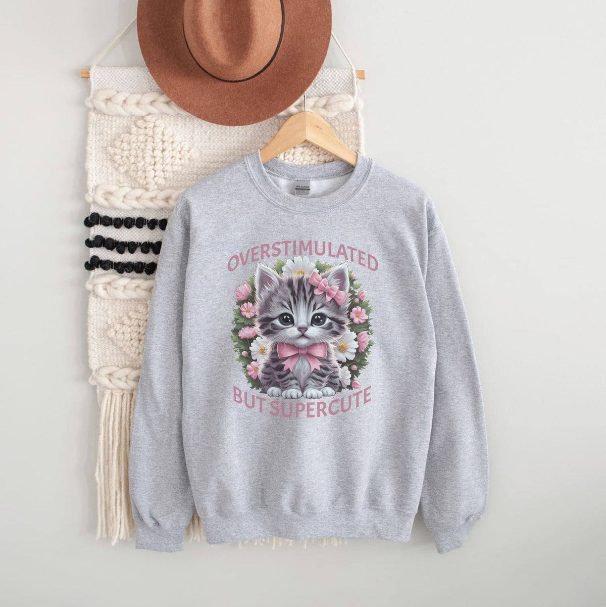 Gray neurospicy sweatshirt with a kitten graphic and text, hanging on a white wall with a hat and decorative items.