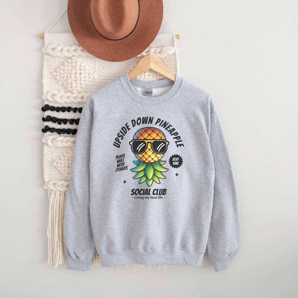 Gray swingers sweatshirt with pineapple graphic and text, hanging on a white wall with a hat and bag.