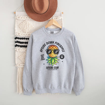 Gray swingers sweatshirt with pineapple graphic and text, hanging on a white wall with a hat and bag.