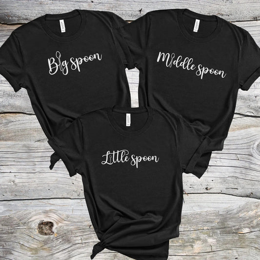 Three matching ENM polyamory black t-shirts with 'Big spoon', 'Middle spoon', and 'Little spoon' text on a wooden surface.