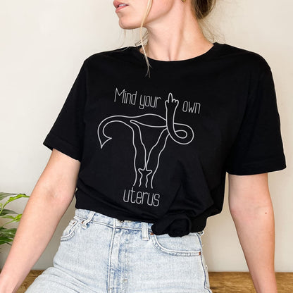 Person wearing a black feminist t-shirt with a graphic and text 'Mind your own uterus'.