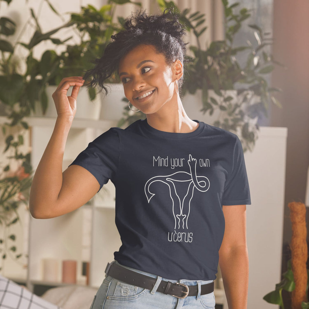 Woman wearing a navy blue t-shirt with a feminist graphic design and text, standing in a room with plants.