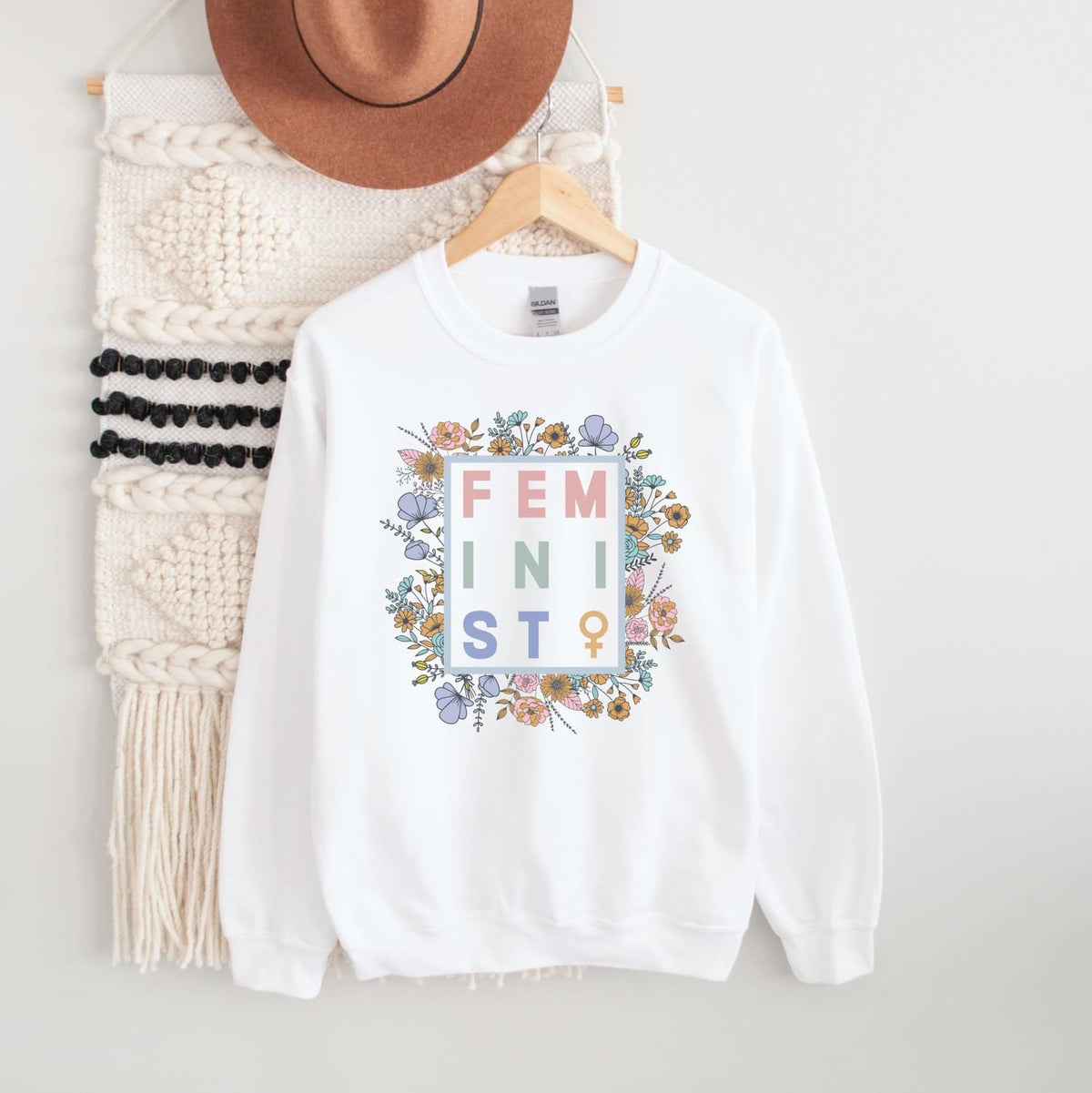 White sweatshirt with 'FEMINIST' text and floral design on a hanger with a hat and scarf.