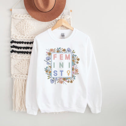 White sweatshirt with 'FEMINIST' text and floral design on a hanger with a hat and scarf.