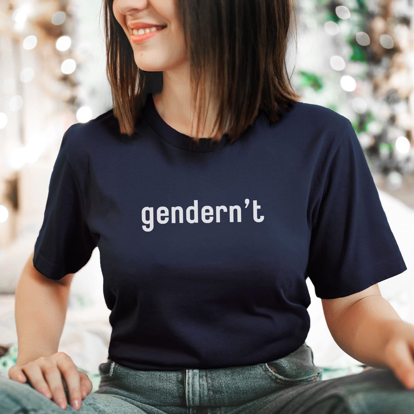 Person wearing a navy blue nonbinary t-shirt with 'gendernt' text, sitting against a blurred indoor background.