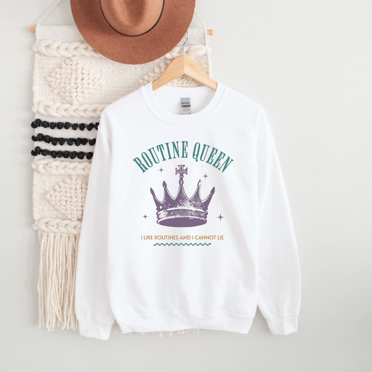 White sweatshirt with 'Routine Queen' text and crown graphic on a hanger with a hat and scarf.