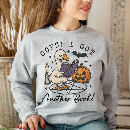 Person wearing a gray sweatshirt with a graphic of a duck holding a book and pumpkin, with text 'Oops! I got another book!'.