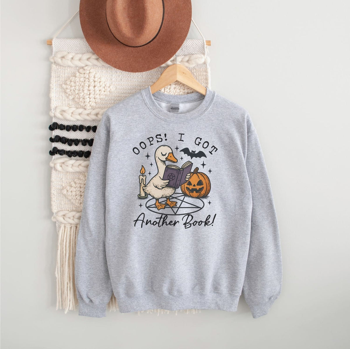 Gray sweatshirt with a graphic design of a duck reading a book next to a pumpkin, with text 'Oops! I got another book!' on a white background.