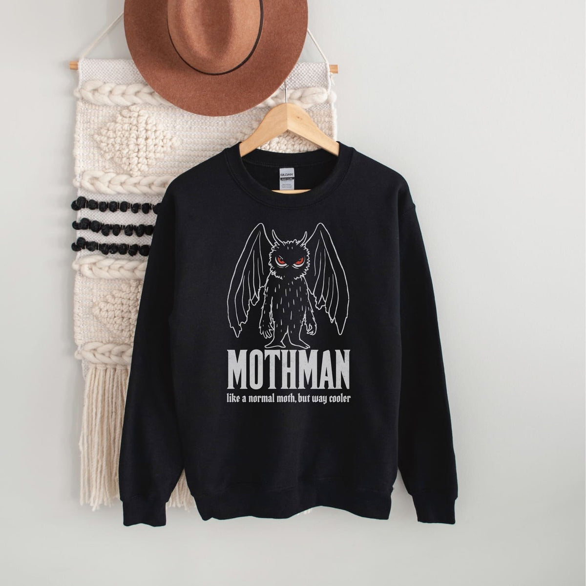 Black sweatshirt with Mothman graphic and text on a hanger against a white wall.