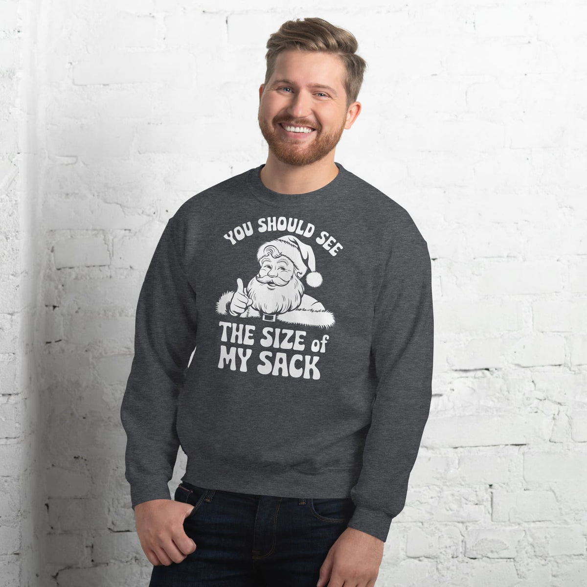Man wearing a dark gray naughty christmas sweatshirt with a graphic and text against a white brick wall.
