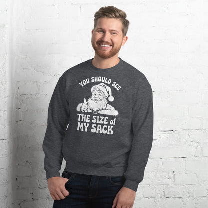 Man wearing a dark gray naughty christmas sweatshirt with a graphic and text against a white brick wall.