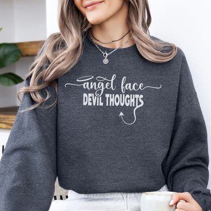Person wearing a dark gray funny naughty sweatshirt with 'angel face devil thoughts' text, holding a mug.