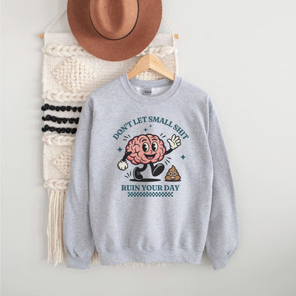 Gray positivity sweatshirt with a cartoon brain design and text, hanging on a white wall with a hat and bag.