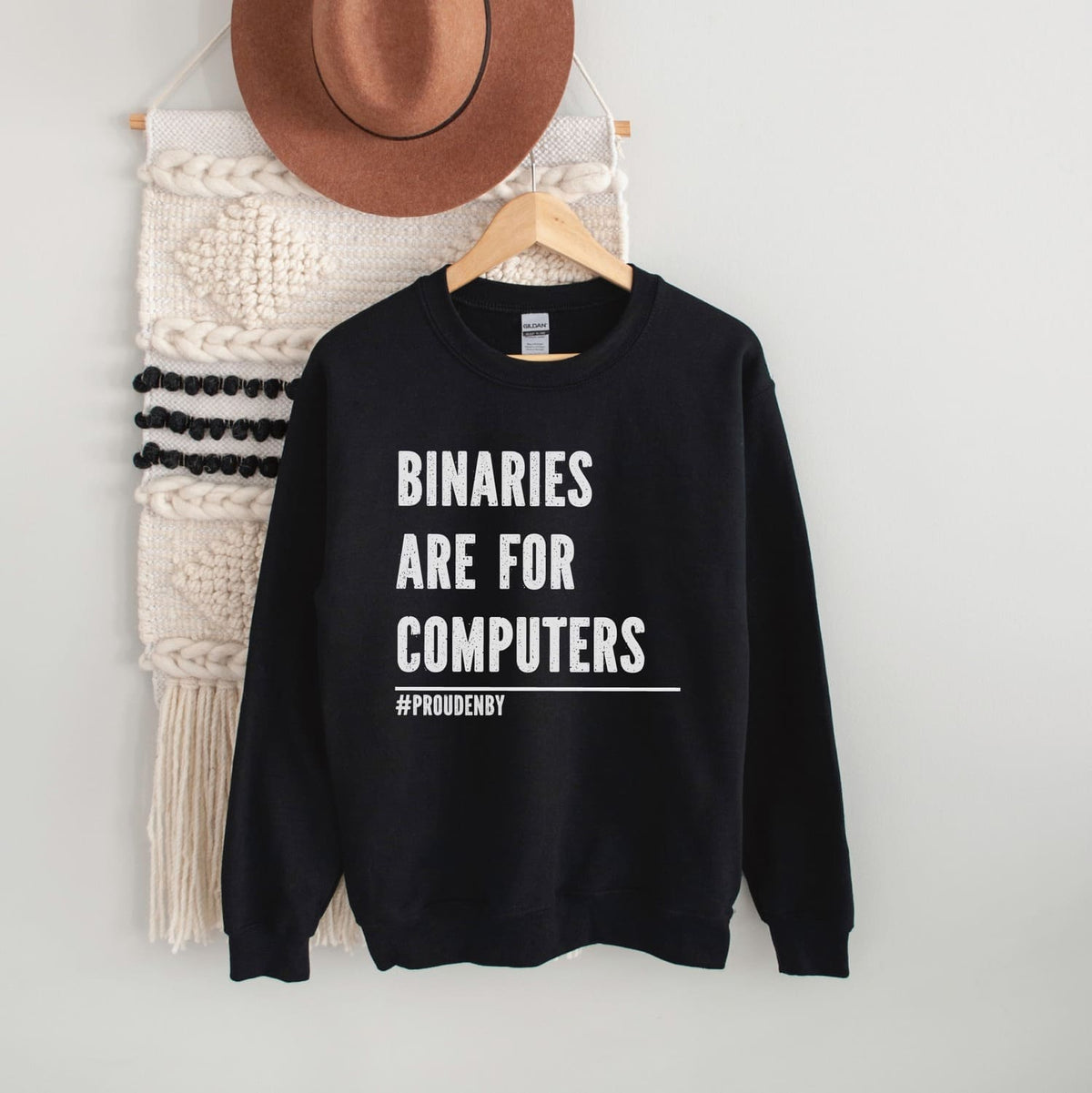 Nonbinary sweatshirt with 'BINARIES ARE FOR COMPUTERS #PROUDENBY' text on a hanger against a white wall.