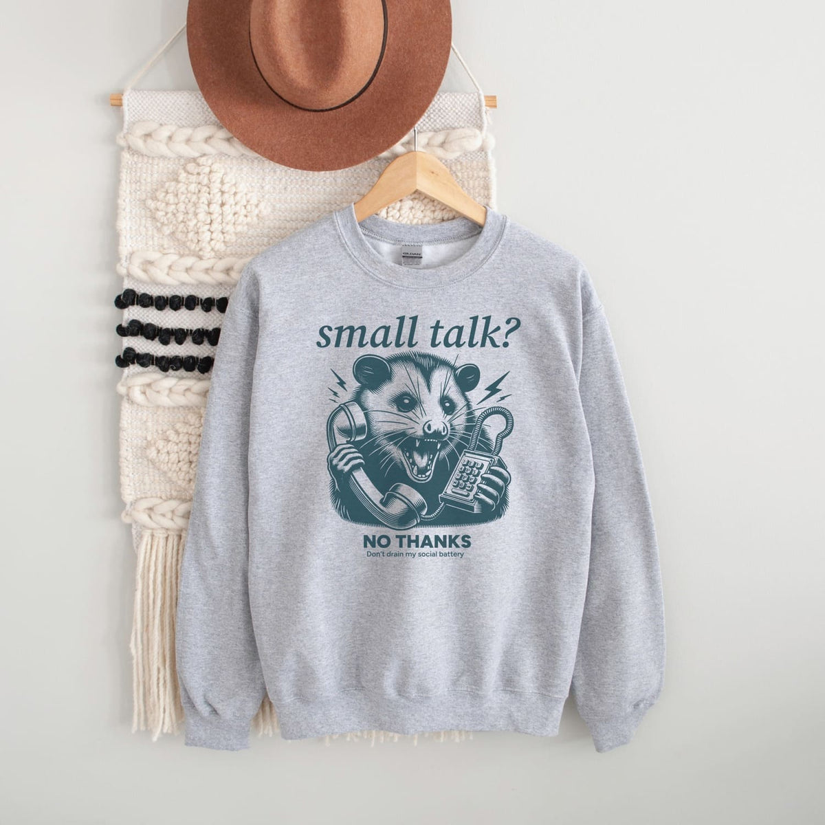 Gray sweatshirt with graphic social anxiety design and text, placed on a white surface with a hat and bag in the background.