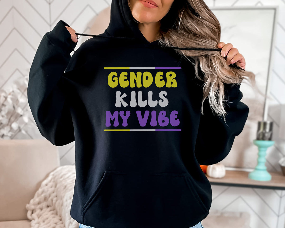 Person wearing a black nonbinary hoodie with colorful text 'GENDER KILLS MY VIBE' in a home setting.