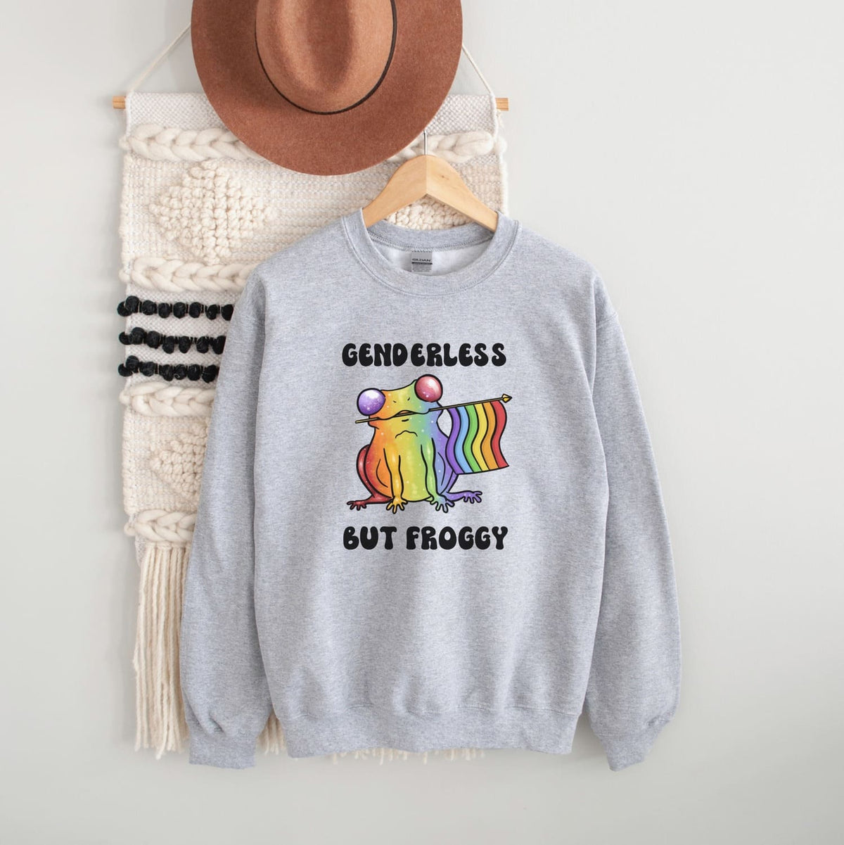 Gray sweatshirt with a rainbow agender or nonbinary frog graphic and text, hanging on a white wall with a hat and bag in the background.