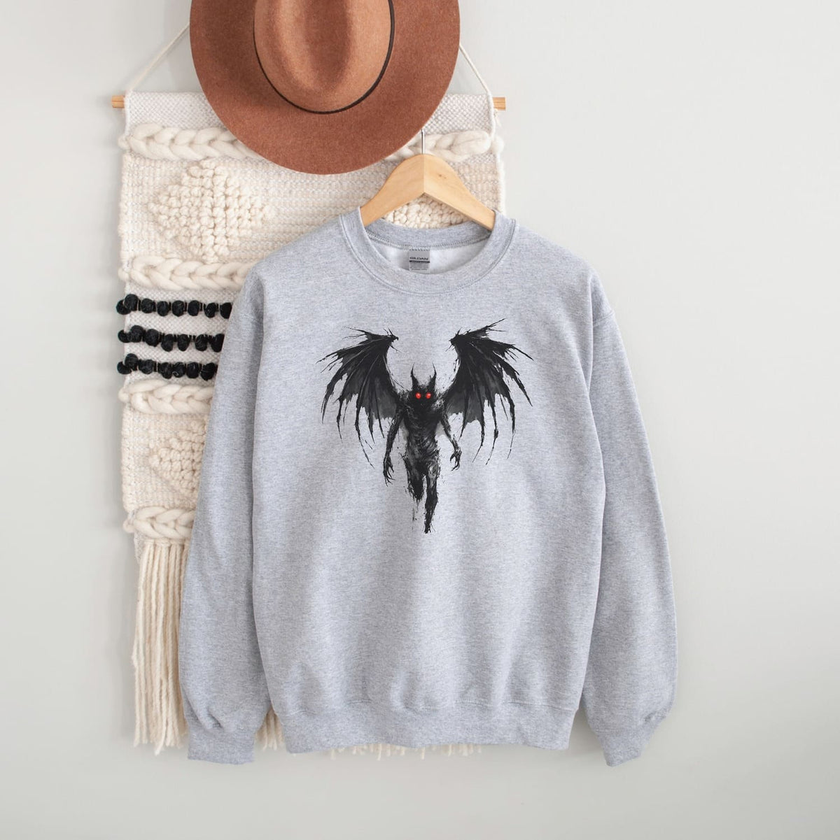 Gray sweatshirt with a mothman design on a hanger against a white wall.