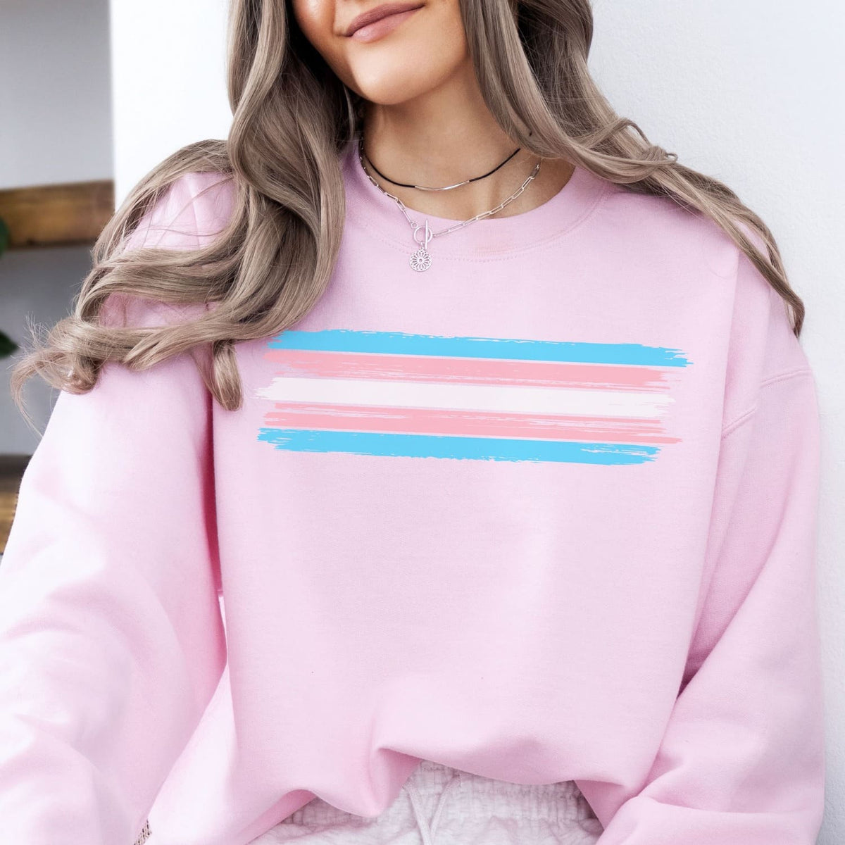 Person wearing a pink trans pride sweatshirt with trangender flag, sitting indoors.